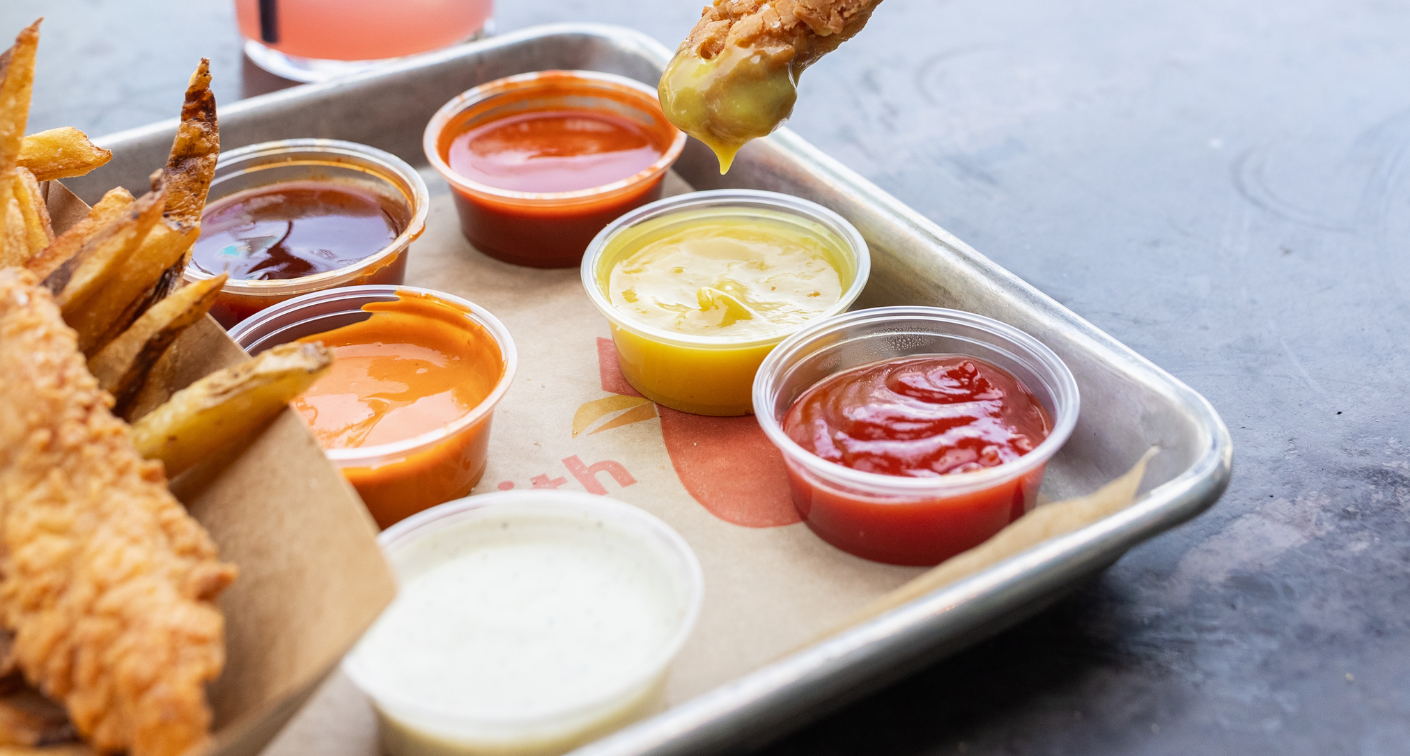 Six different sauces on a tray next to chicken tenders.